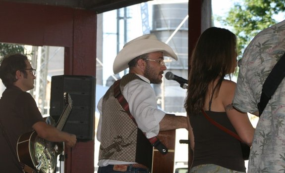 Outdoor music scene: a bearded man in a white hat sings into a microphone beside a guitarist as people listen.